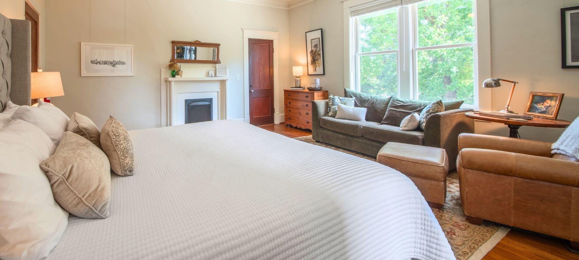 Guest room at The Gillen House Bed and Breakfast with a king bed, fireplace, sofa, leather chair, hardwood floors, and large windows overlooking trees.