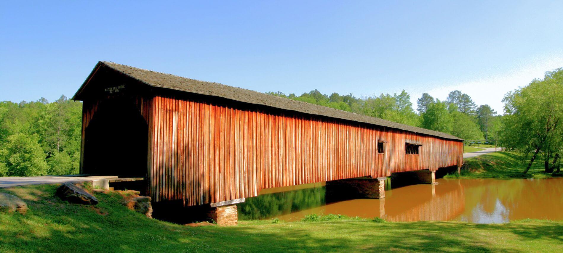 The Watson Mill Covered Bridge over a calm river with grassy banks and trees on a sunny day.